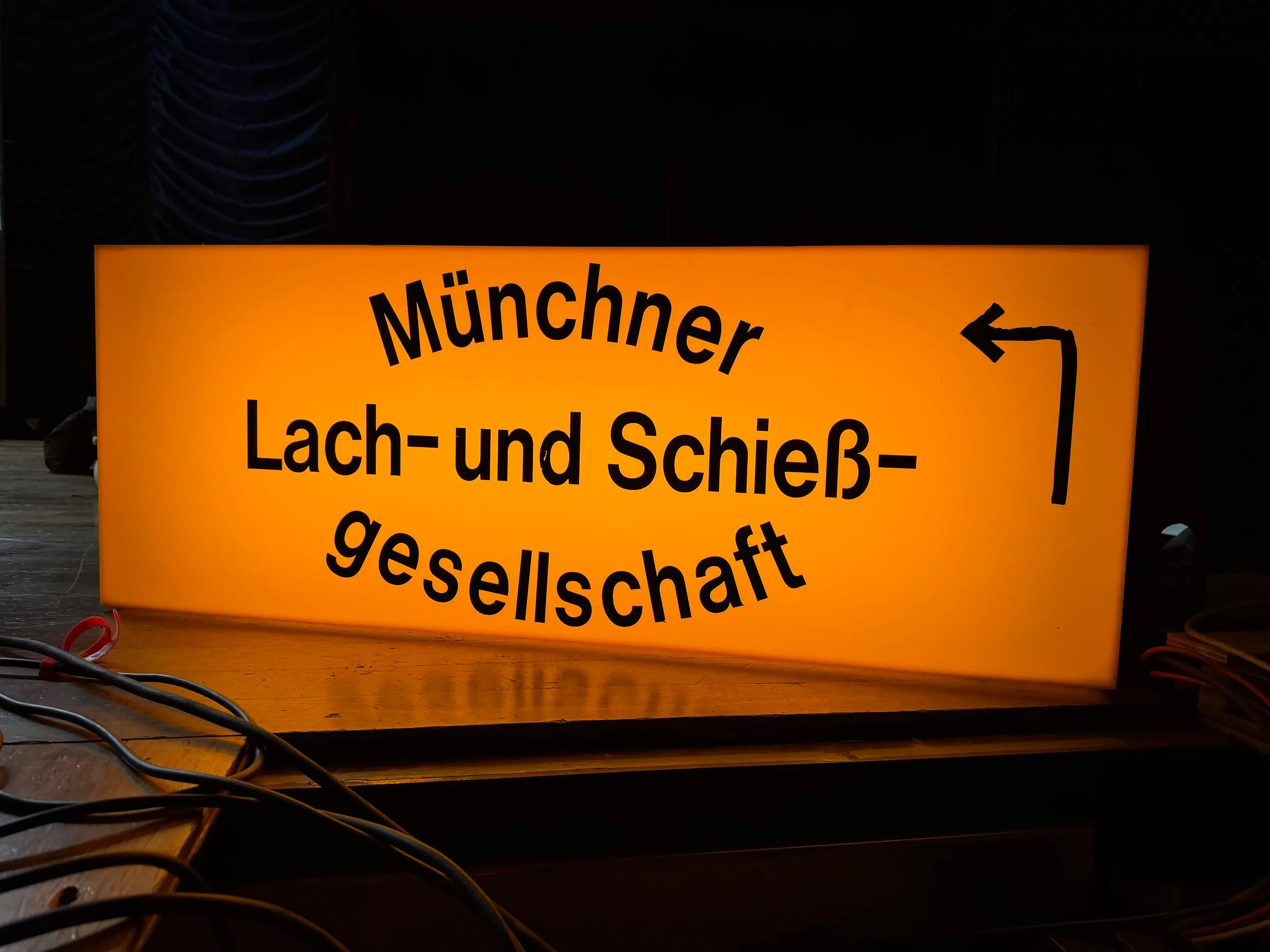 Leuchtschild „Münchner Lach- und Schießgesellschaft“ mit Richtungspfeil