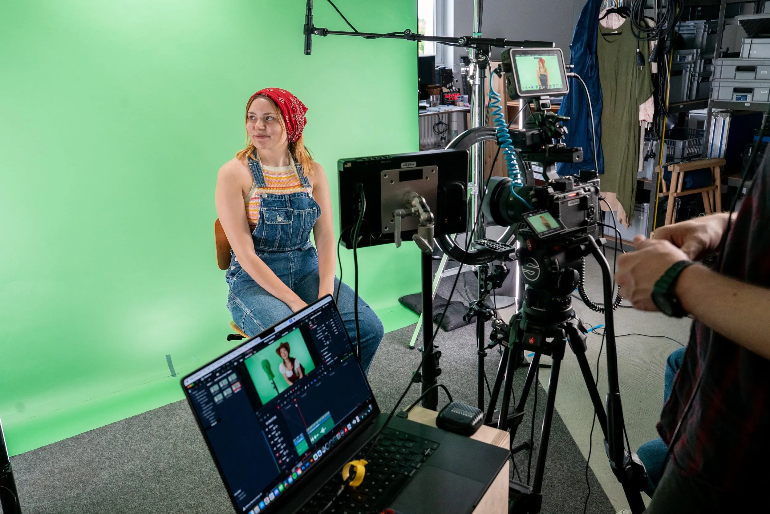 Darstellerin mit rotem Kopftuch sitzt vor dem Greenscreen, umgeben von Kamera, Monitor und Laptop