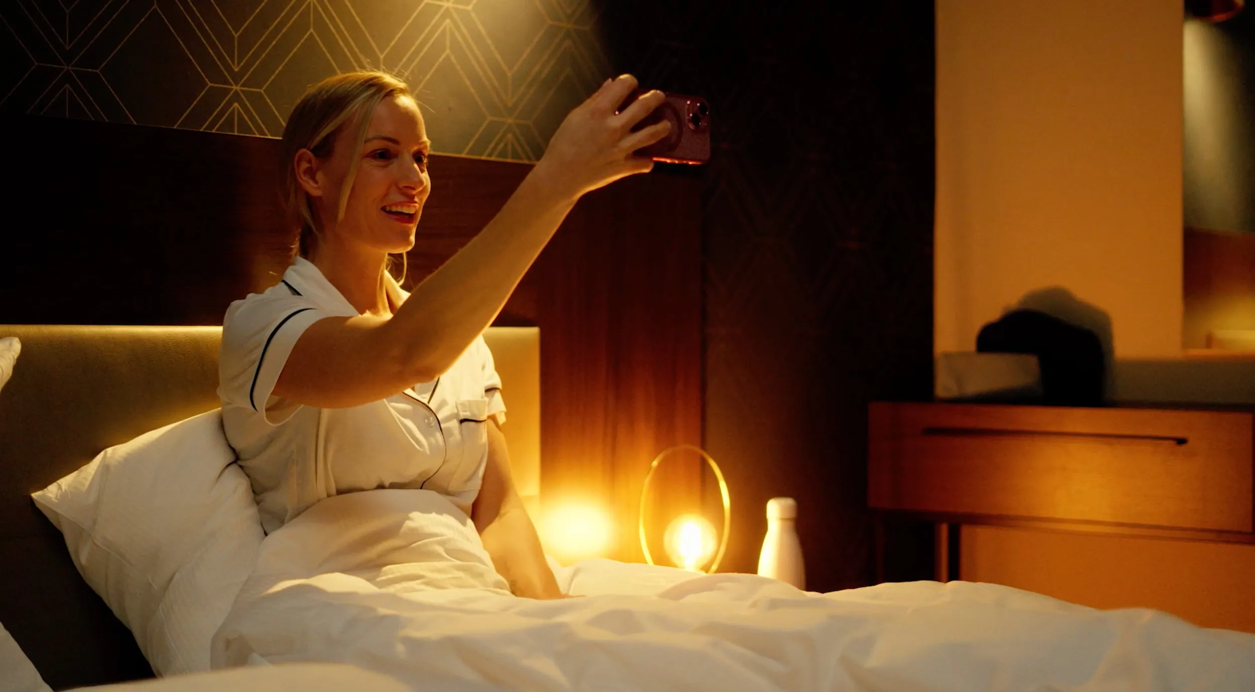 Frau macht im Hotelbett bei warmem Lampenlicht ein Selfie