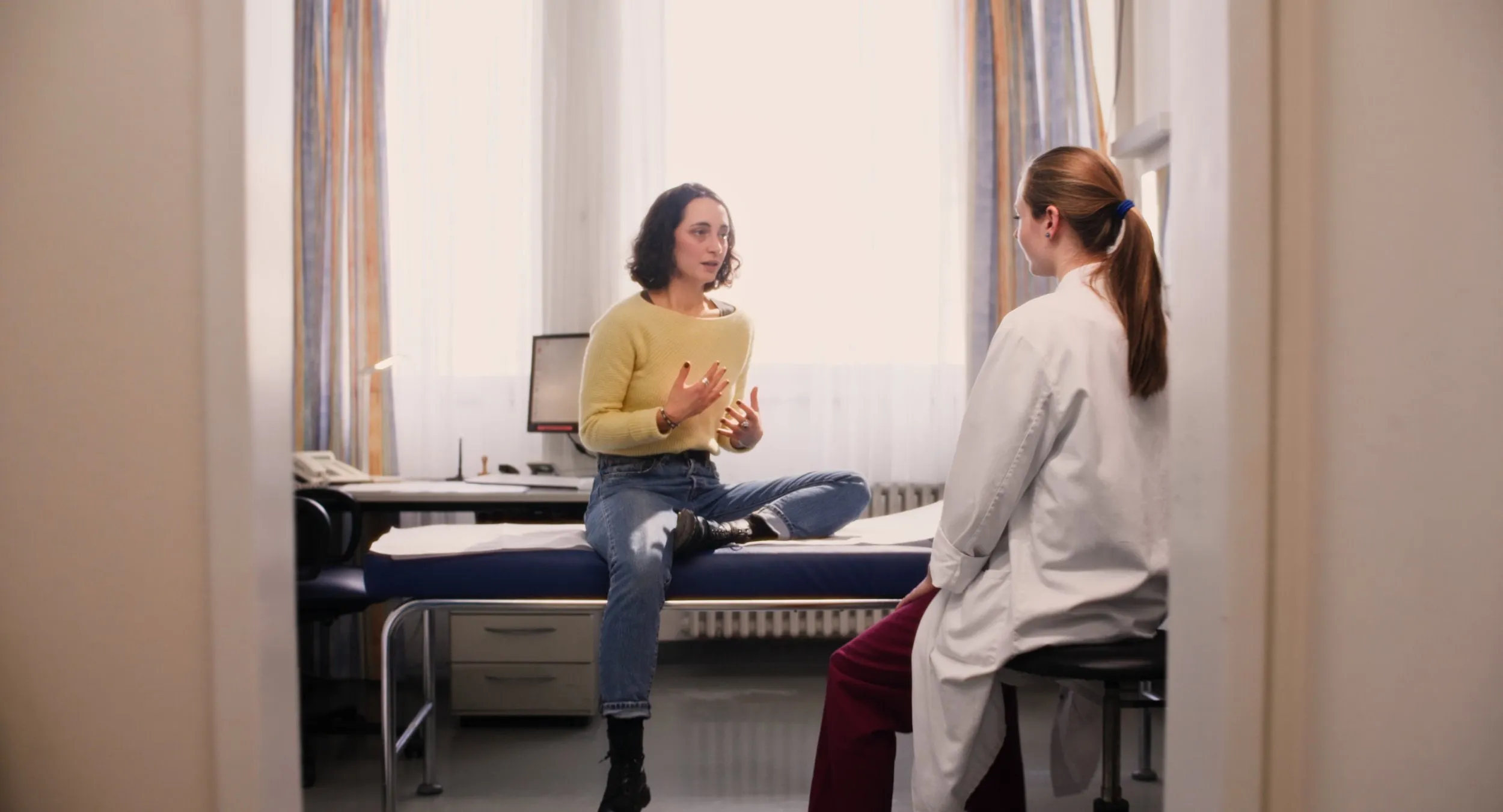 Patientin im gelben Pullover im Gespräch mit einer Ärztin im Untersuchungszimmer