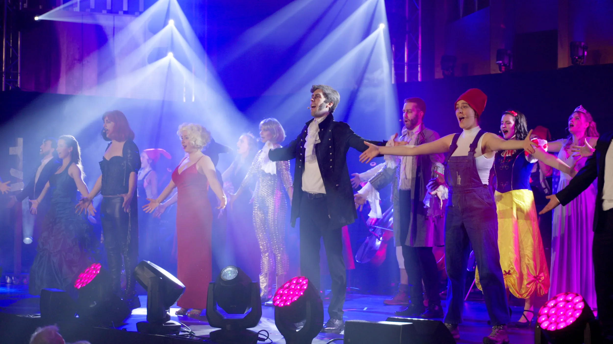 Ensemble in Kostümen auf der Bühne im blauen und pinken Scheinwerferlicht