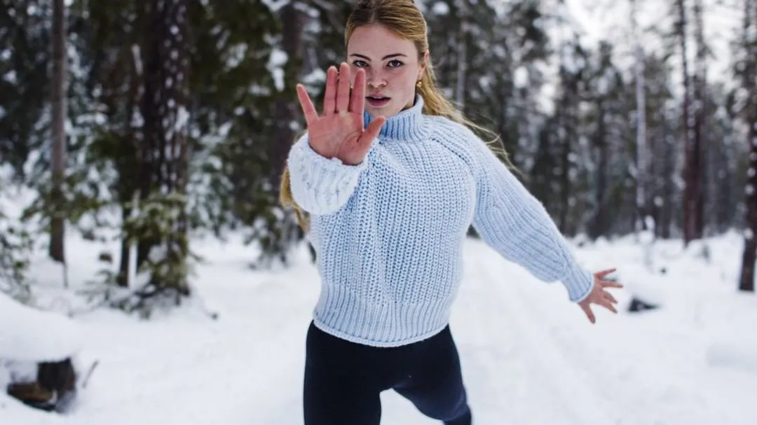 Eiskunstläuferin im weißen Strickpullover streckt im verschneiten Wald eine Hand zur Kamera