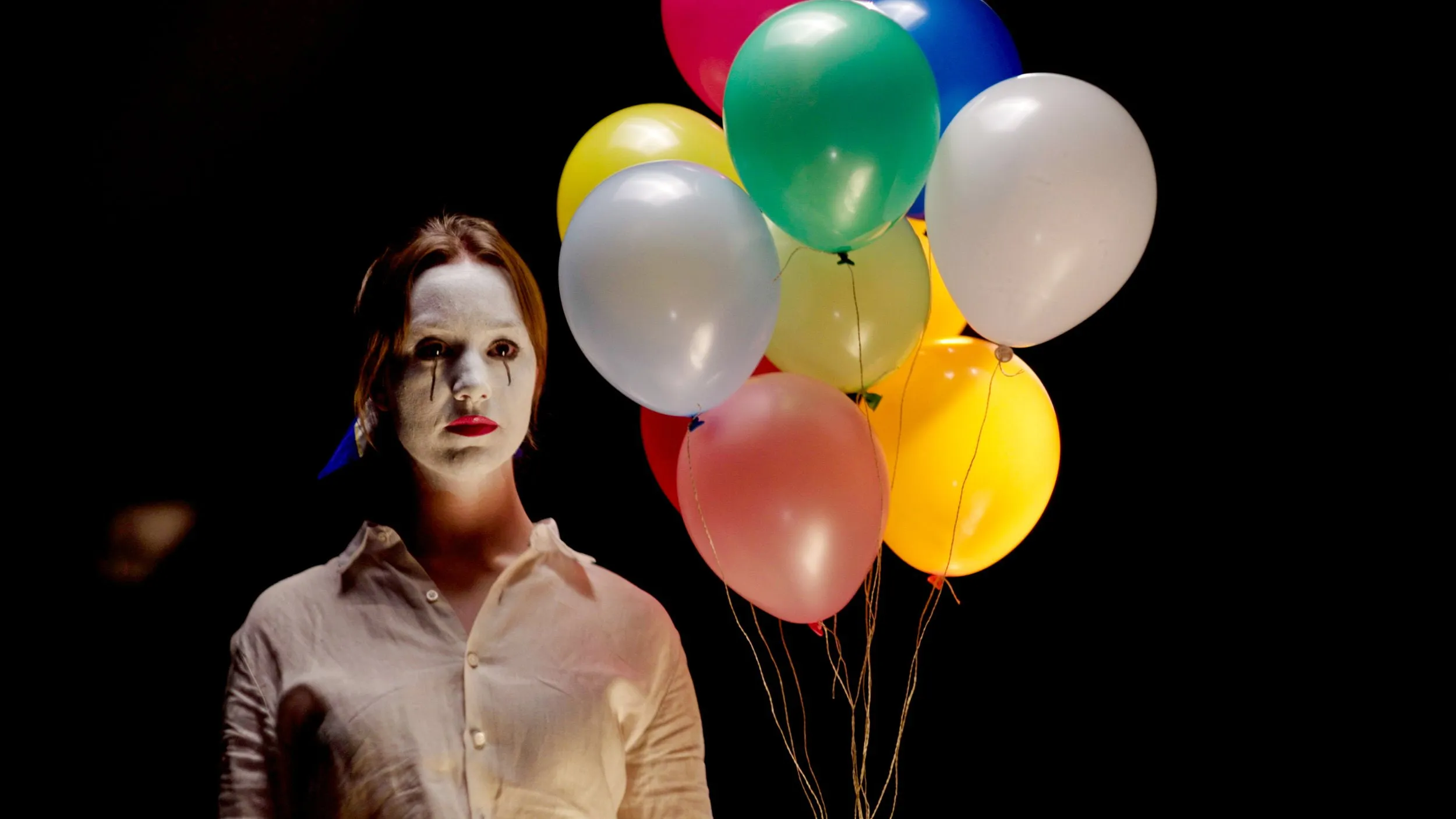 Darstellerin mit weiß geschminktem Gesicht hält bunte Luftballons vor schwarzem Hintergrund