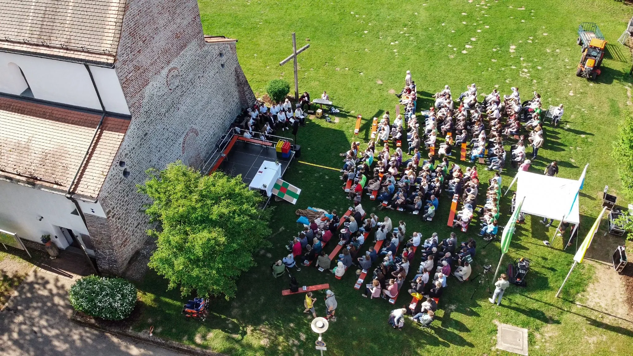 Luftaufnahme eines Open-Air-Gottesdienstes neben einer Kirche mit vielen Teilnehmenden auf einer Wiese