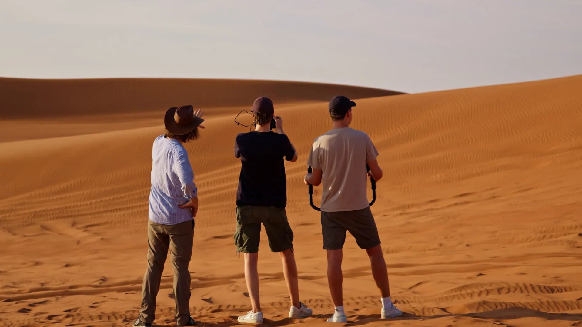 Drei Filmemacher mit Kameraausrüstung stehen von hinten gesehen in der Wüste und blicken über die Dünen
