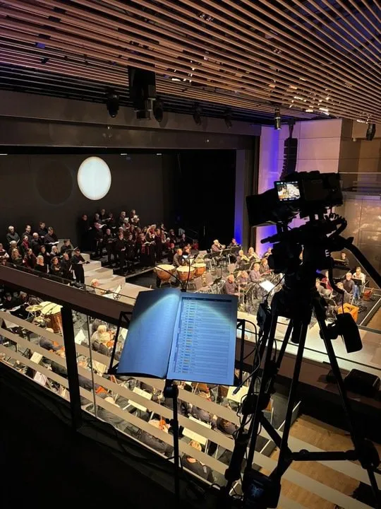 Kamera auf Stativ mit Partitur auf einer Empore über einem Konzertsaal mit Orchester